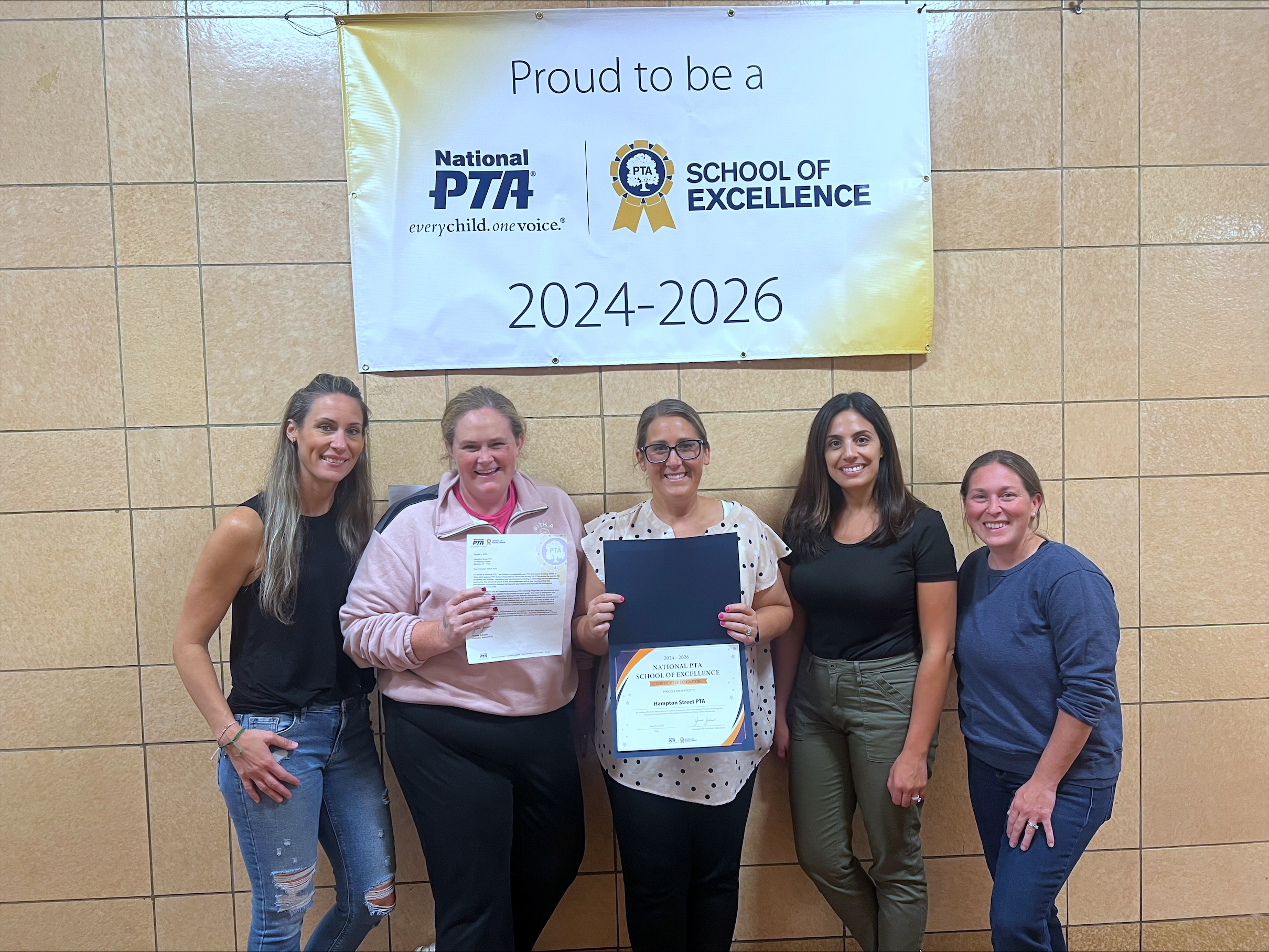 Five women smiling under a school of excellence banner with one holding a certificate